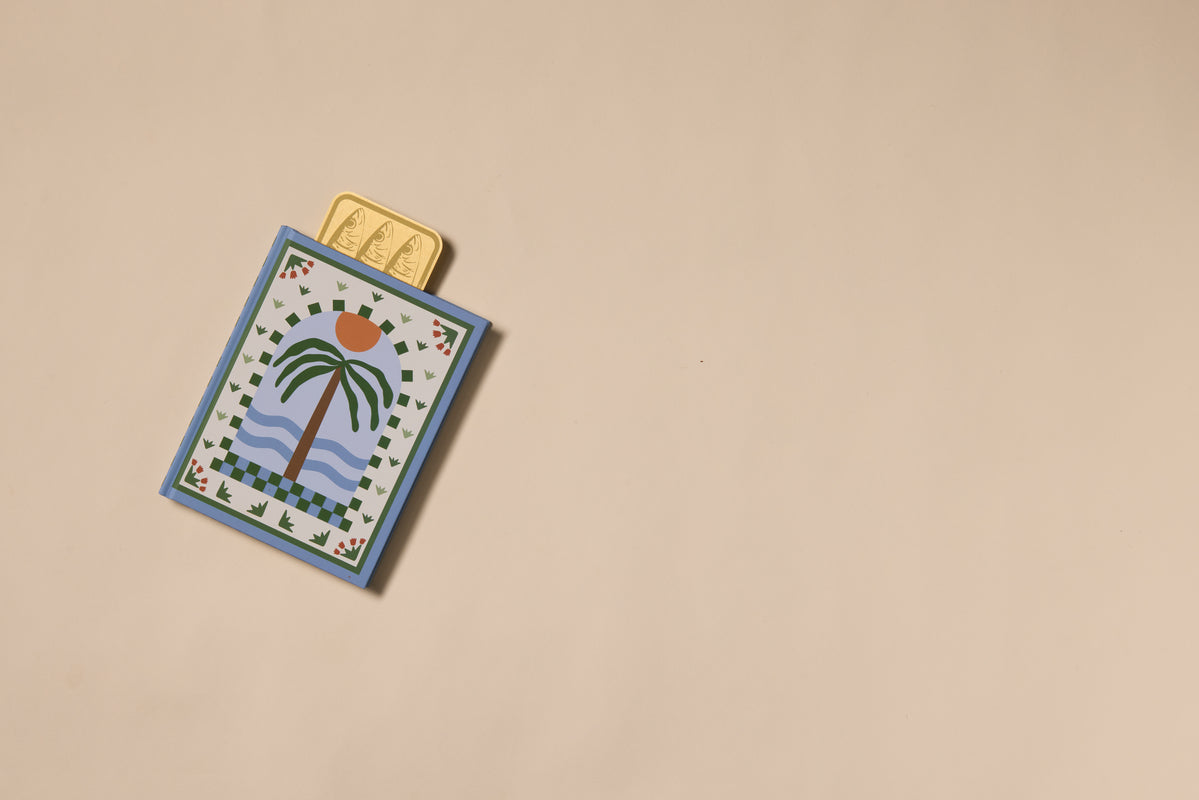 A decorative card with a palm tree and sunset design lies on a beige surface. Three Brass Bookmark - Sardines, featuring golden sardine motifs, are partially tucked inside the card against a plain, uncluttered background.
