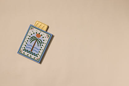 A decorative card with a palm tree and sunset design lies on a beige surface. Three Brass Bookmark - Sardines, featuring golden sardine motifs, are partially tucked inside the card against a plain, uncluttered background.
