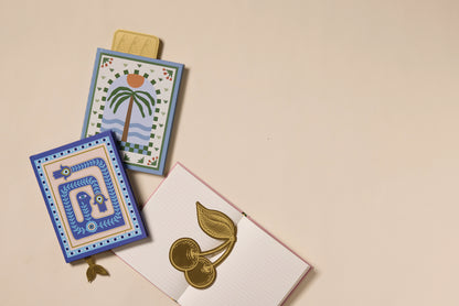 Two decorative notepads—one with a palm tree and sun, the other with blue patterns—are placed beside an open lined notebook adorned with the Brass Bookmark - Cherries, all set on a light beige surface.