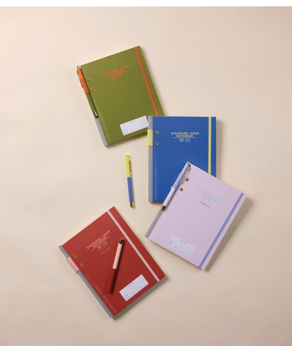 Four colorful notebooks (green, blue, red, and pink) with lined labels and gold ring bindings are arranged on a beige surface, each paired with a Color Block Pen in Rosewood + Blush placed diagonally across or beside them.