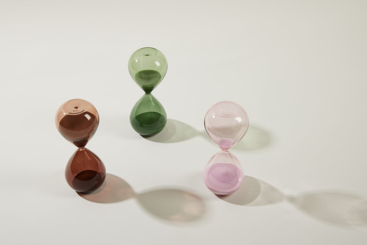 The Hourglass, 15 min - Evergreen stands elegantly on a white surface as a meditation timer, its green sand casting a soft, elongated shadow to the right.