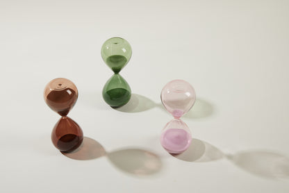 The Hourglass, 15 min - Evergreen stands elegantly on a white surface as a meditation timer, its green sand casting a soft, elongated shadow to the right.