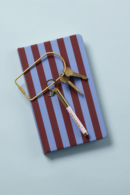 A Soft Touch Journal in Purple + Nutmeg Stripe rests on a light blue surface, topped with a gold key ring holding two keys and a gold IMPORTANT-labeled pen.