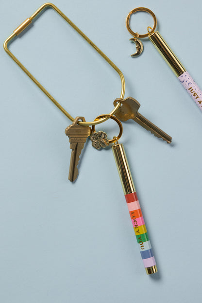 A gold key ring with two keys, a four-leaf clover charm, and the Pen Keychain - Lucky You (a colorful compact ink pen with "LUCKY YOU" on it). Another gold key ring holds a crescent moon charm and a pink keychain pen on the blue background.