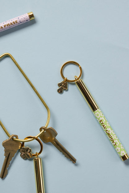 The Pen Keychain - Stay Groovy is a compact floral ink pen on a gold key ring with a clover charm, displayed on a light blue background.