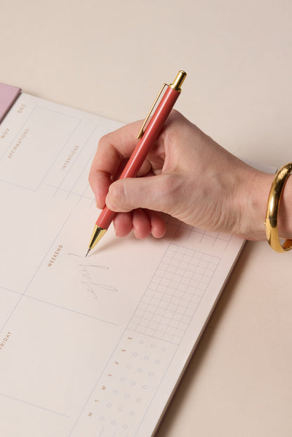 A hand wearing a gold bracelet writes “yay” on a weekly planner using the Rosewood + Blush Mechanical Pencil, perfect for colorful planning or neat notes with its 0.7mm lead, on a pale-background planner featuring calendar and grid sections.