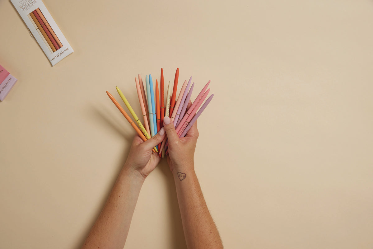 Two hands display a fan of Slim Metal Twisty Pens - Confidence Set on a beige background, with a heart tattoo on one wrist and two boxes of the inspirational pens visible in the top left corner.