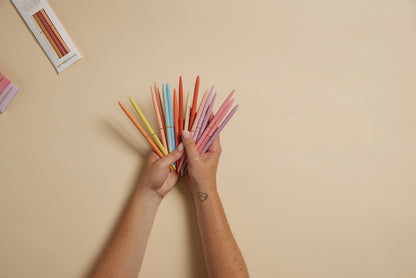 Two hands display a fan of Slim Metal Twisty Pens - Confidence Set on a beige background, with a heart tattoo on one wrist and two boxes of the inspirational pens visible in the top left corner.