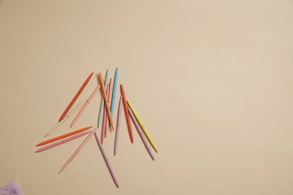 A set of Slim Metal Twisty Pens - Motivation, featuring black ink in pink, orange, blue, yellow, and peach colors, is scattered on a light beige surface with plenty of empty space to the right.