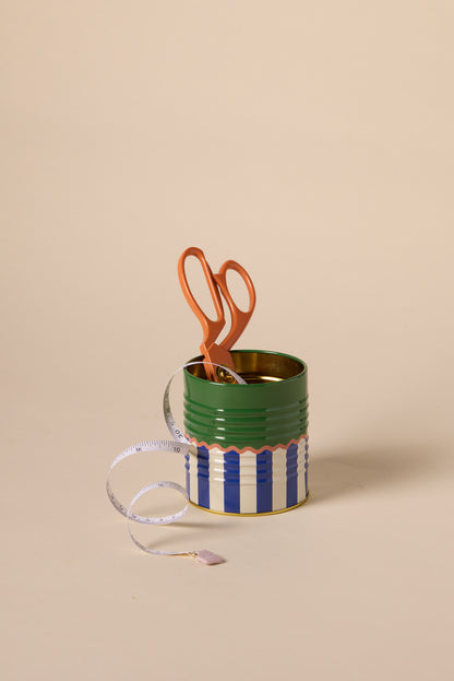 A pair of orange scissors stands upright in the A Dopo Solid & Striped Storage Tin, with a white measuring tape featuring a lavender end draped over the front, all set against a plain beige background.