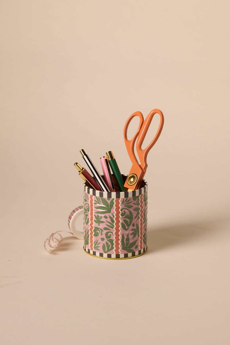 The A Dopo Jungle Storage Tin, featuring a vibrant pink and green pattern, holds pens, pencils, and large orange scissors—ideal for your desk or as a fun alternative to a keepsake box. Curled ribbon accents the tin on a light beige background.