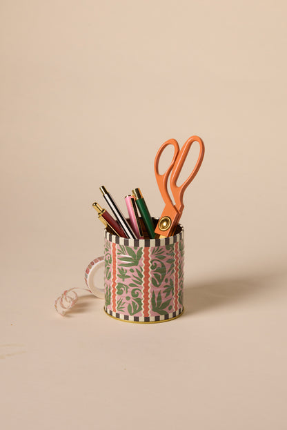 The A Dopo Jungle Storage Tin, featuring a vibrant pink and green pattern, holds pens, pencils, and large orange scissors—ideal for your desk or as a fun alternative to a keepsake box. Curled ribbon accents the tin on a light beige background.