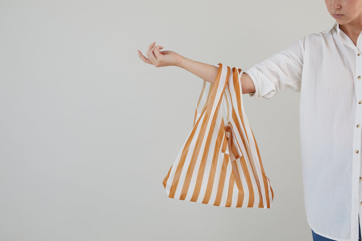 A person wearing a white button-up shirt holds the Reusable Market Bag - Stripe, featuring yellow-and-white stripes, on their extended arm against a plain light background.
