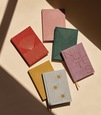 Six Hard Cover Suede Cloth Journals With Pocket—each with "NOTES" in bold, featuring geometric designs in red, pink, green, yellow, blue, and lavender—are arranged on a beige surface with striking diagonal shadows.