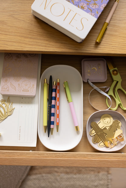 A wooden drawer contains Color Block Pens - Lilac + Cornflower with ultra fine ballpoint tips on a white tray, gold scissors, decorative paper clips, a measuring tape, a notepad, and other stationery items neatly organized.