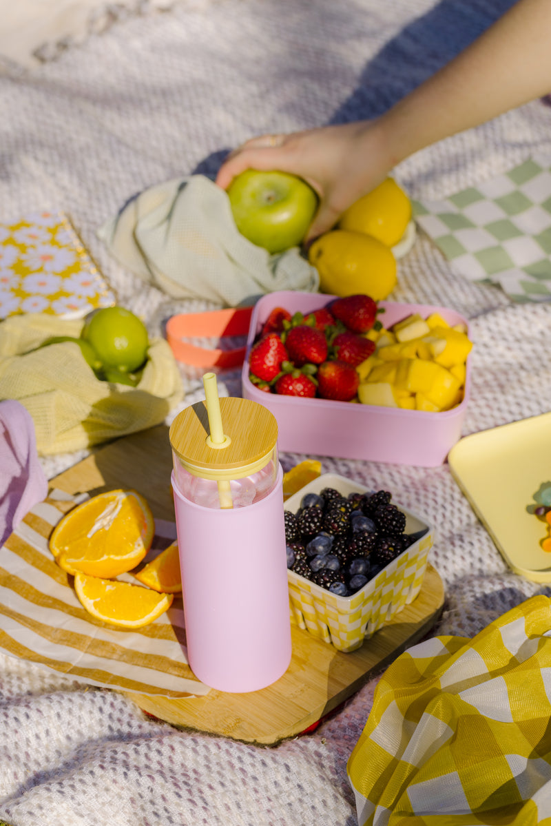 A picnic set on a blanket features the Tumbler With Straw - Citron/Pink, containers of strawberries, mango, blackberries, sliced oranges, a green apple, a lime, and a hand reaching for an apple in a bright sunny outdoor scene.