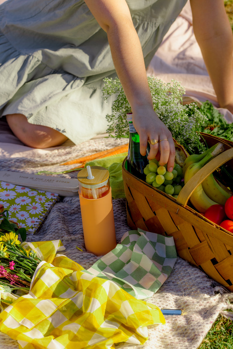 A person in a light green dress sits on a picnic blanket, reaching into a wicker basket of grapes, bananas, tomatoes, and flowers. Nearby are napkins, wildflowers, and the Tumbler With Straw - Mint/Peach for sipping.