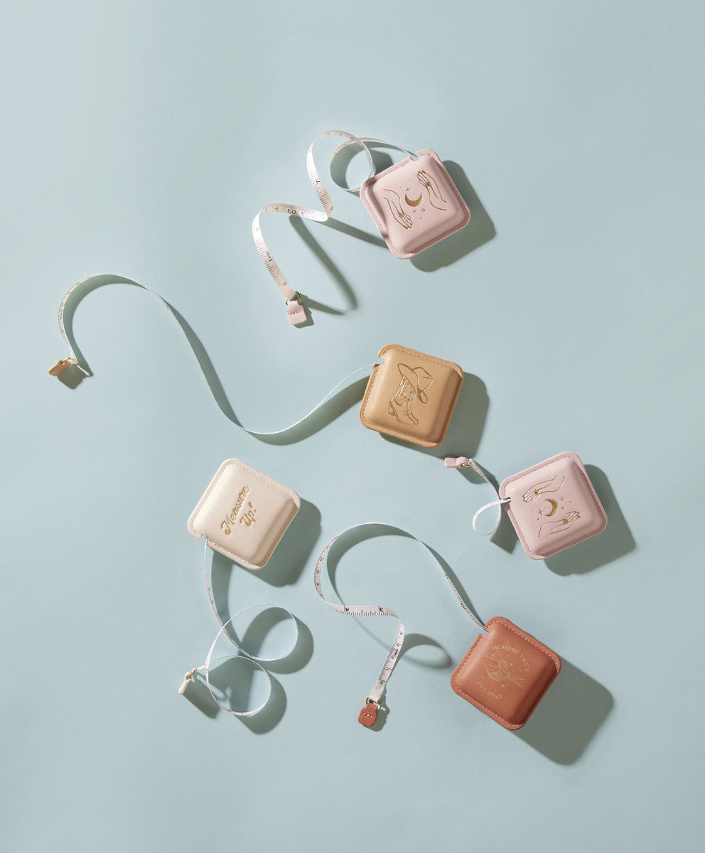 Five Pocket Tape Measure - Measure Twice in pastel cases—pink, beige, tan—are arranged on a light blue surface. Each vegan leather tape measure has a unique gold illustration, with tapes partially extended and curled around them.