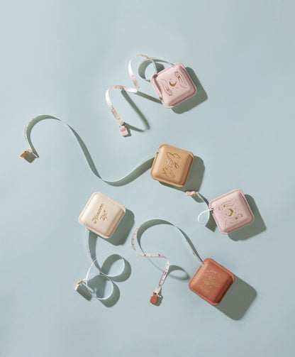 Five Pocket Tape Measure - Measure Twice in pastel cases—pink, beige, tan—are arranged on a light blue surface. Each vegan leather tape measure has a unique gold illustration, with tapes partially extended and curled around them.