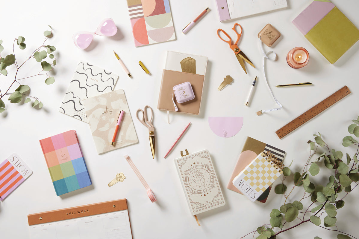 A flat lay on a white background features colorful notebooks, planners, pens, scissors, the Pocket Tape Measure - Cowgirl with a stylish design, washi tape, a ruler, a candle, and leafy branches. The stationery showcases pastel colors and various patterns.