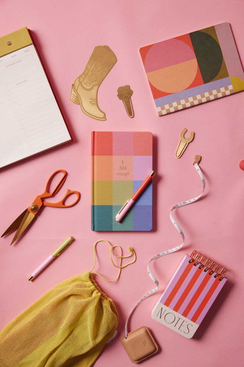 Colorful stationery, such as a striped notebook, planner, pens, scissors, the "Pocket Tape Measure - Measure Twice," sticky notes, and gold paper clips in fun shapes are arranged on a pink background.