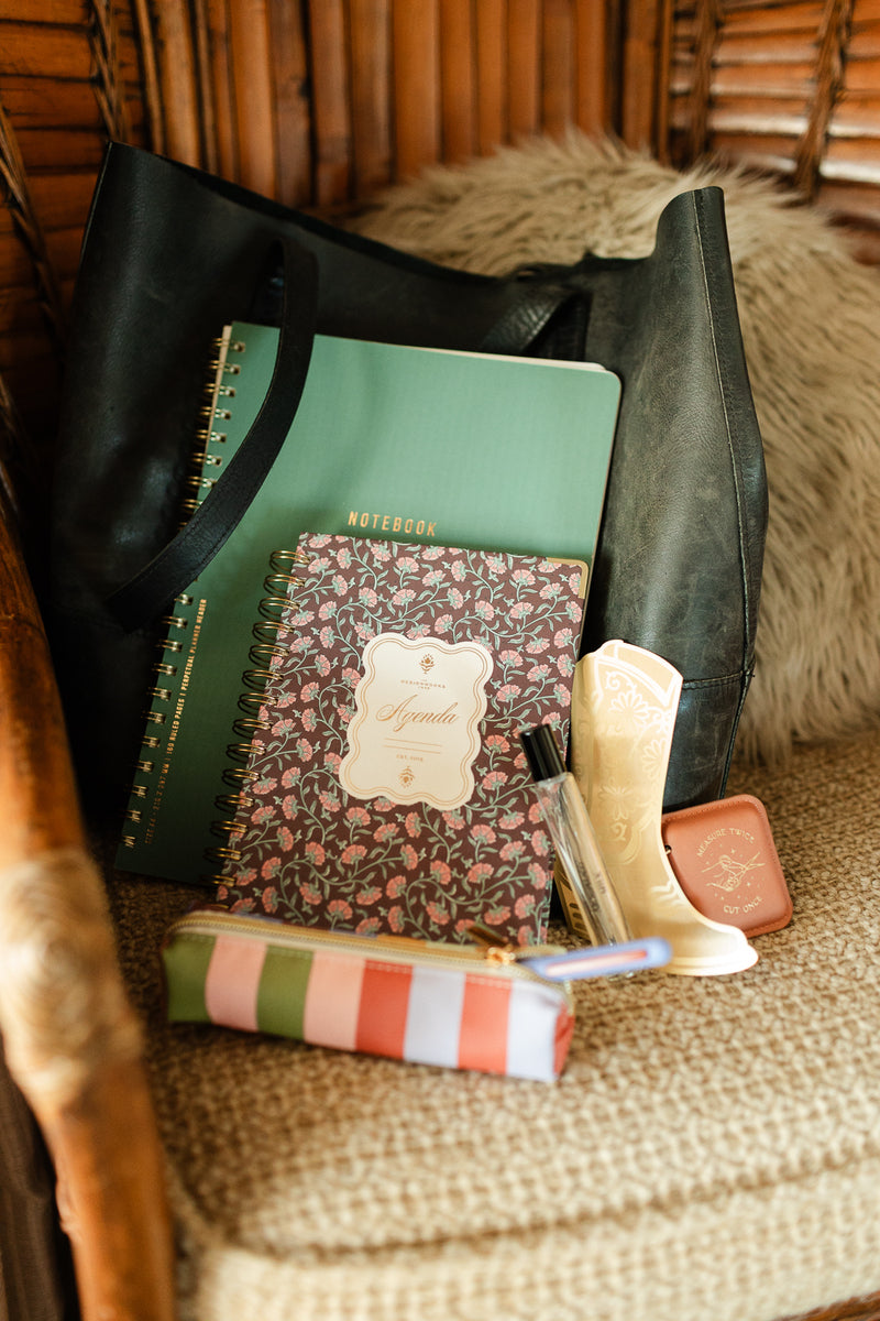 A black tote bag on a cushioned chair holds a green notebook, a floral agenda, the Pen & Pouch Set - Bold Stripe, a perfume bottle, a decorative boot-shaped item, and a compact mirror.