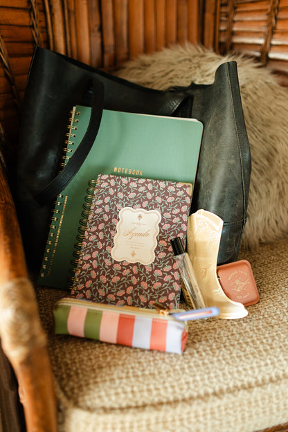 A black tote bag on a cushioned chair holds a green notebook, a floral agenda, the Pen & Pouch Set - Bold Stripe, a perfume bottle, a decorative boot-shaped item, and a compact mirror.
