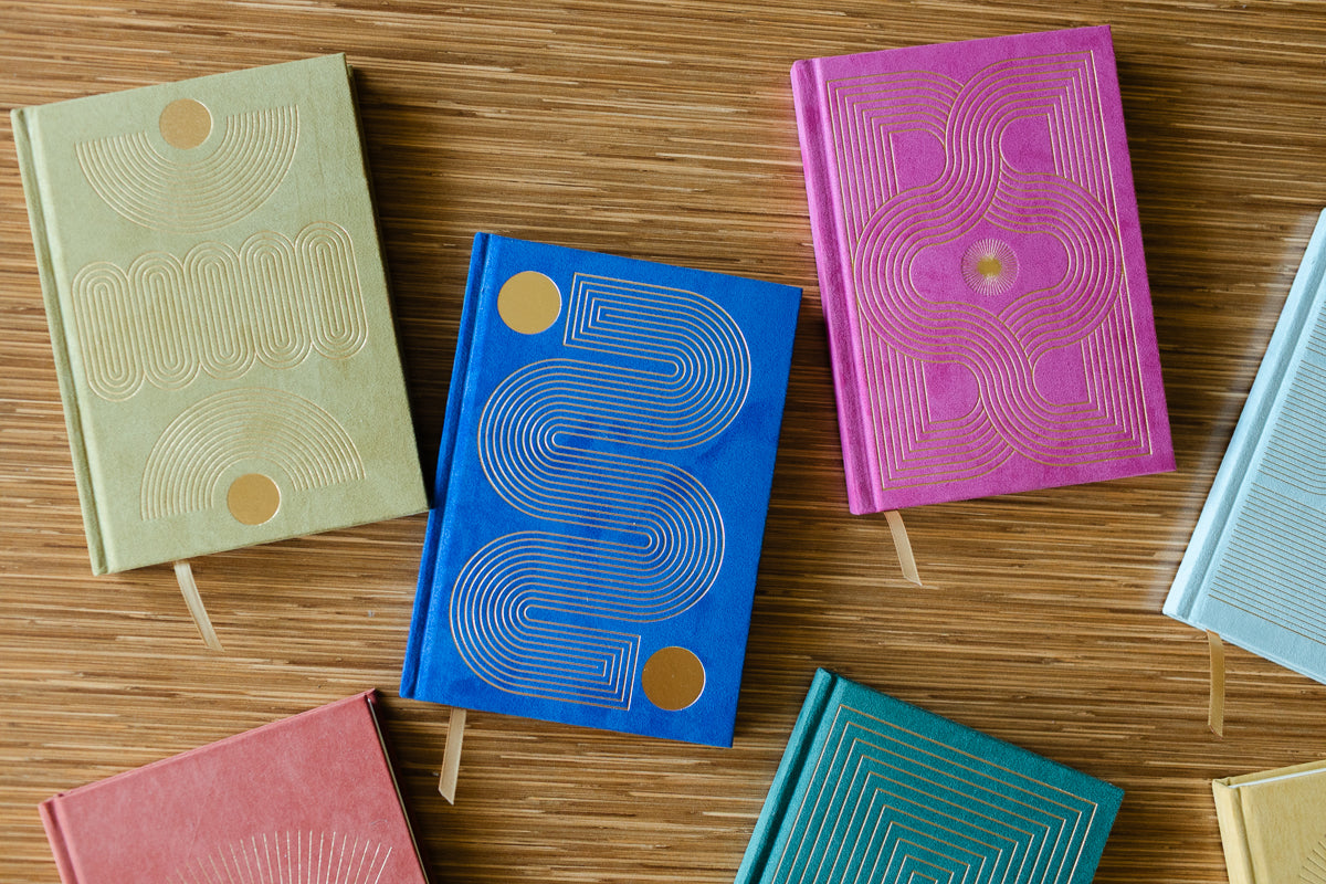 The Radiant Journal - Fuchsia, a colorful hardcover with lined pages and a ribbon bookmark, is featured alongside eco-friendly journals with geometric designs and gold foil accents, all arranged on a wooden surface.