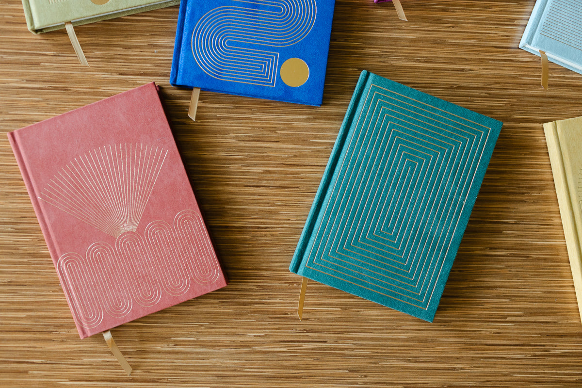 Radiant Journal - Rays features colorful hard covers with geometric gold designs and ribbon bookmarks, available in blue, red, green, and cream. Each journal contains lined pages for organized notes or reflections.