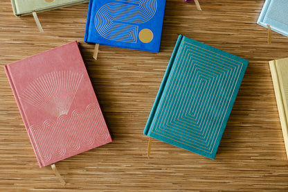 Radiant Journal - Rays features colorful hard covers with geometric gold designs and ribbon bookmarks, available in blue, red, green, and cream. Each journal contains lined pages for organized notes or reflections.