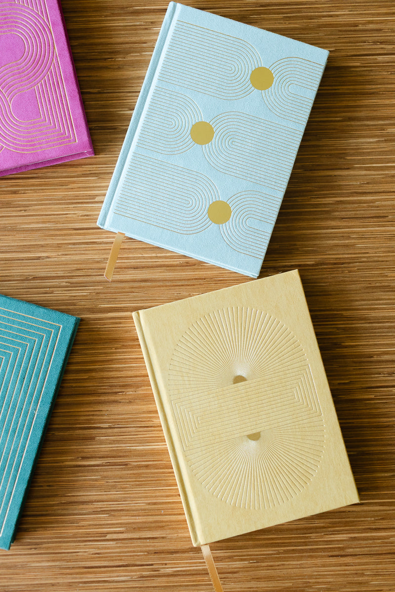Four Hard Cover Suede Cloth Journals With Pocket—Arch Dot designs in light blue, yellow, pink, and teal—rest on a wooden surface. Each lined notebook features geometric embossing and a ribbon bookmark for easy organization.