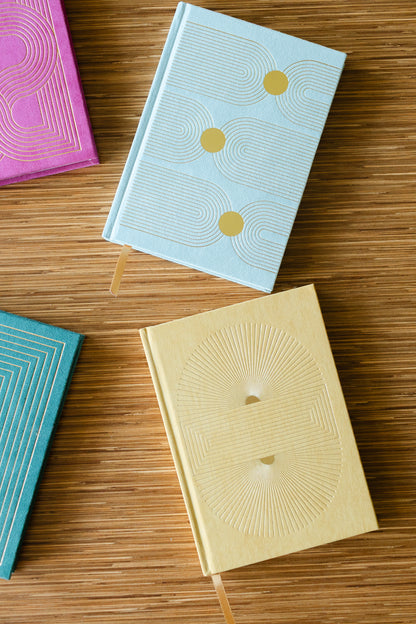 Four Hard Cover Suede Cloth Journals With Pocket—Arch Dot designs in light blue, yellow, pink, and teal—rest on a wooden surface. Each lined notebook features geometric embossing and a ribbon bookmark for easy organization.