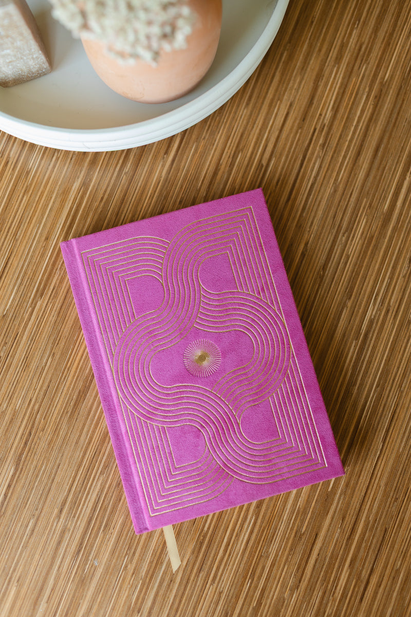 The Radiant Journal - Fuchsia, featuring suede and an intricate gold geometric design, rests on a wooden surface beside a white tray with a potted plant.