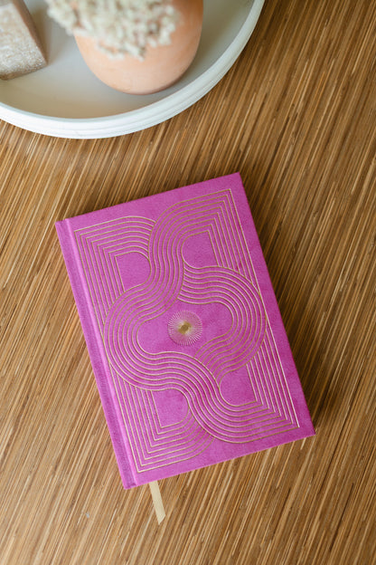 The Radiant Journal - Fuchsia, featuring suede and an intricate gold geometric design, rests on a wooden surface beside a white tray with a potted plant.