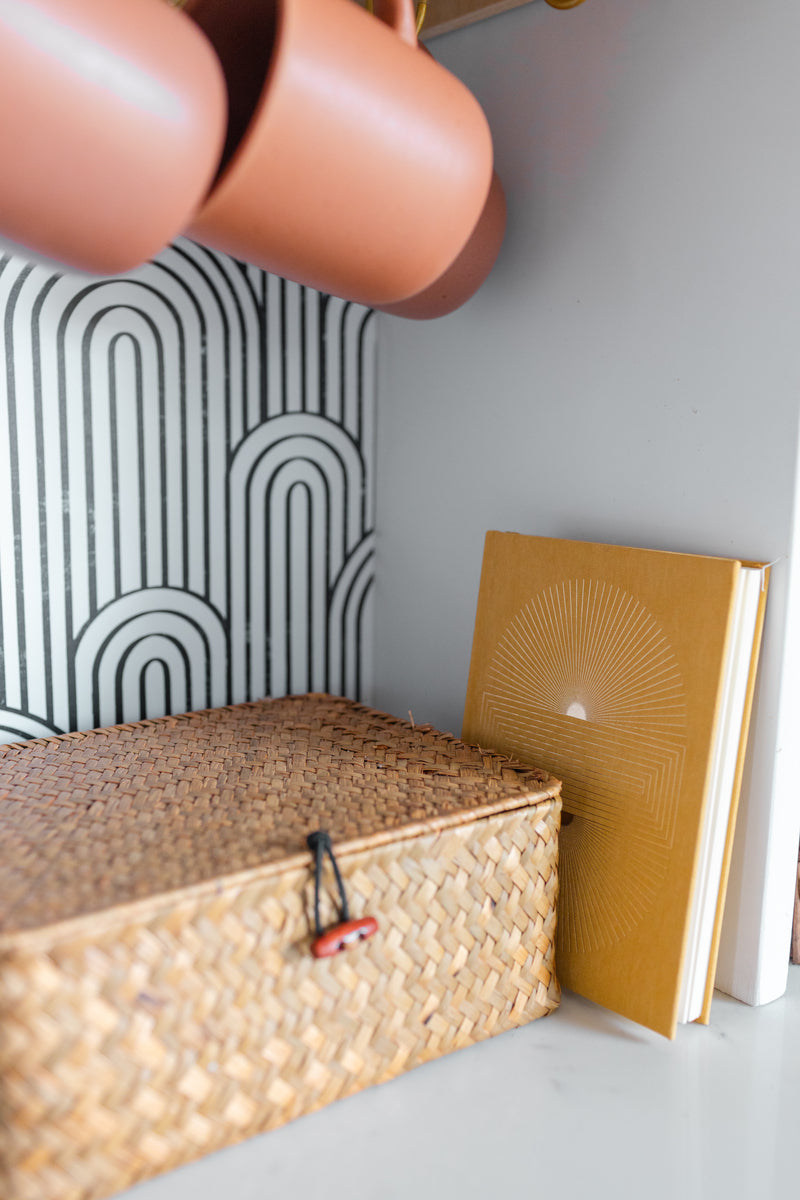 A Radiant Journal - Suns Block with a sunburst design rests in a woven wicker box on a white shelf. Above, terracotta cups hang against a black and white geometric patterned wall.