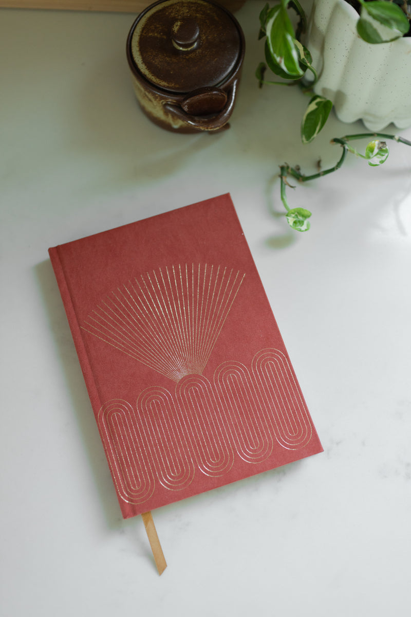 The Radiant Journal - Rays, featuring a red hard cover with gold geometric designs, sits on a white surface next to a brown ceramic jar and a white pot holding a trailing green plant.
