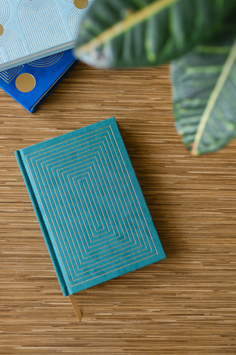 A Hard Cover Suede Cloth Journal With Pocket - Linear Boxes, featuring teal suede with gold geometric lines and a ribbon bookmark, rests on wood. A plant leaf is seen at the top right, with two more journals stacked in the background.