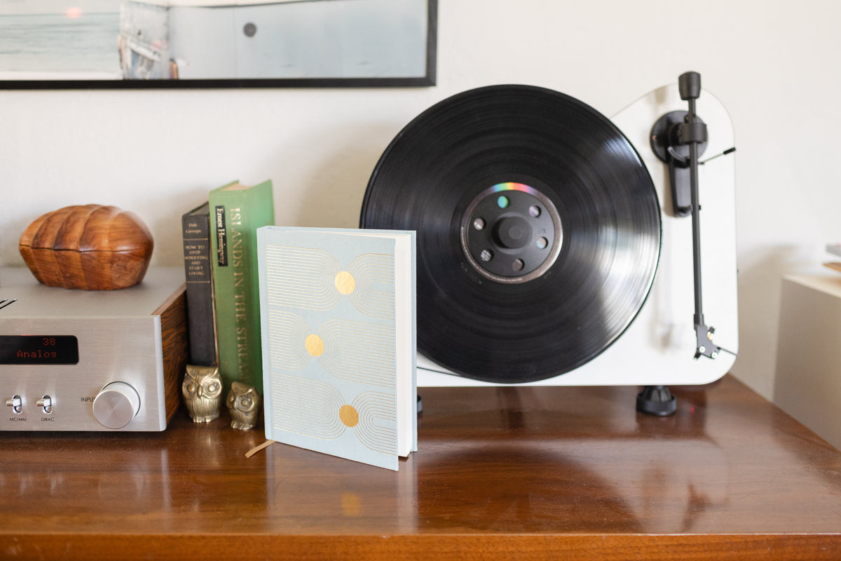 A white record player with a vinyl sits on a wooden table next to books, a stereo, two owl figurines, and the Hard Cover Suede Cloth Journal With Pocket - Arch Dot.