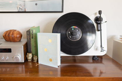 A white record player with a vinyl sits on a wooden table next to books, a stereo, two owl figurines, and the Hard Cover Suede Cloth Journal With Pocket - Arch Dot.