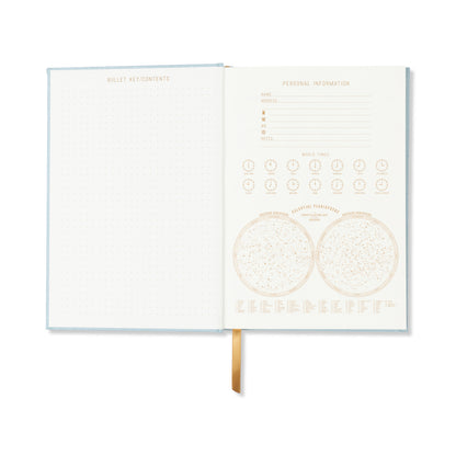 The Hard Cover Suede Cloth Journal With Pocket - Arch Dot in blue showcases a dotted page and a personal info page with world times, celestial pointers, and contact details. A gold ribbon bookmark adds elegance to this journal's stylish design.