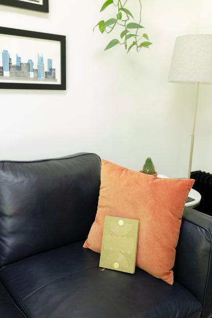 A modern living room with a dark blue leather sofa, an orange cushion holding the closed Radiant Journal - Avocado, plus a potted plant, framed cityscape art, and a floor lamp in the background.
