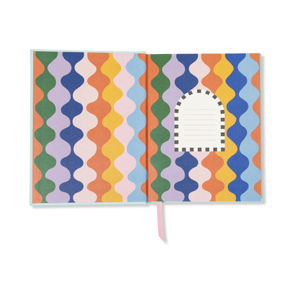 The A Dopo Blue Cheetah Journal is an open, eco-friendly journal featuring wavy, colorful vertical stripes and a small arched white label with lines for writing on the right page. A pink ribbon bookmark peeks out from the bottom, while its lay-flat binding ensures it’s easy to write on every page.