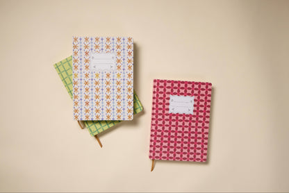Three patterned notebooks, including the Tulip Jumbo Journal - Marigold, are arranged on a beige surface. Each features a ribbon bookmark, geometric patterns in green, orange, and pink, and a white label on the cover.