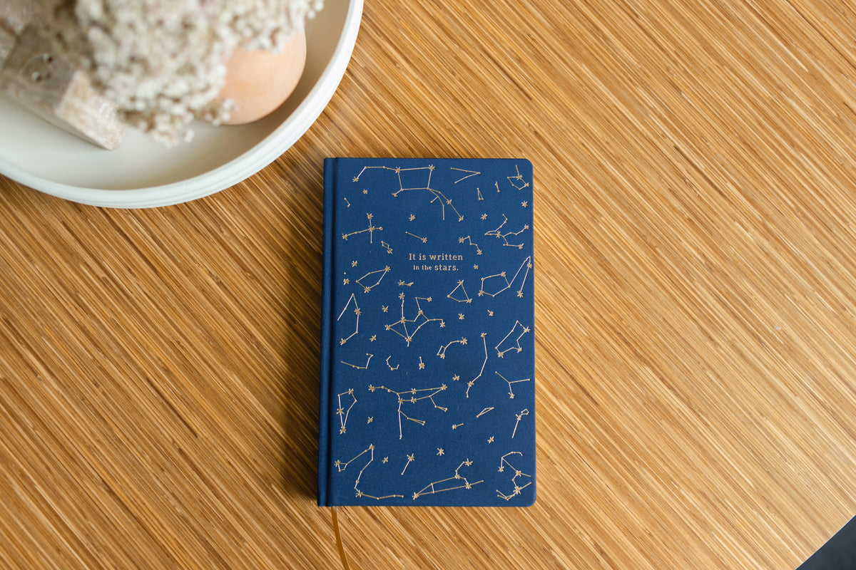 The Cloth Journal - Navy Constellations "It Is Written In The Stars," with gold constellations and lined, lay-flat pages, sits on a wooden table beside a white dish holding a plant.