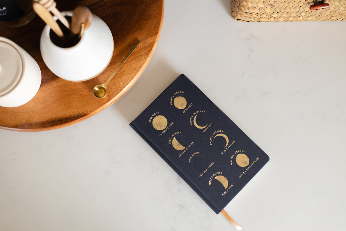 The Cloth Journal - Moon Phases features a navy blue cover with gold moon phases and their names. Shown on a white surface next to a wooden tray with a white vase, utensils, and gold spoon. Includes acid-free paper for lasting notes.