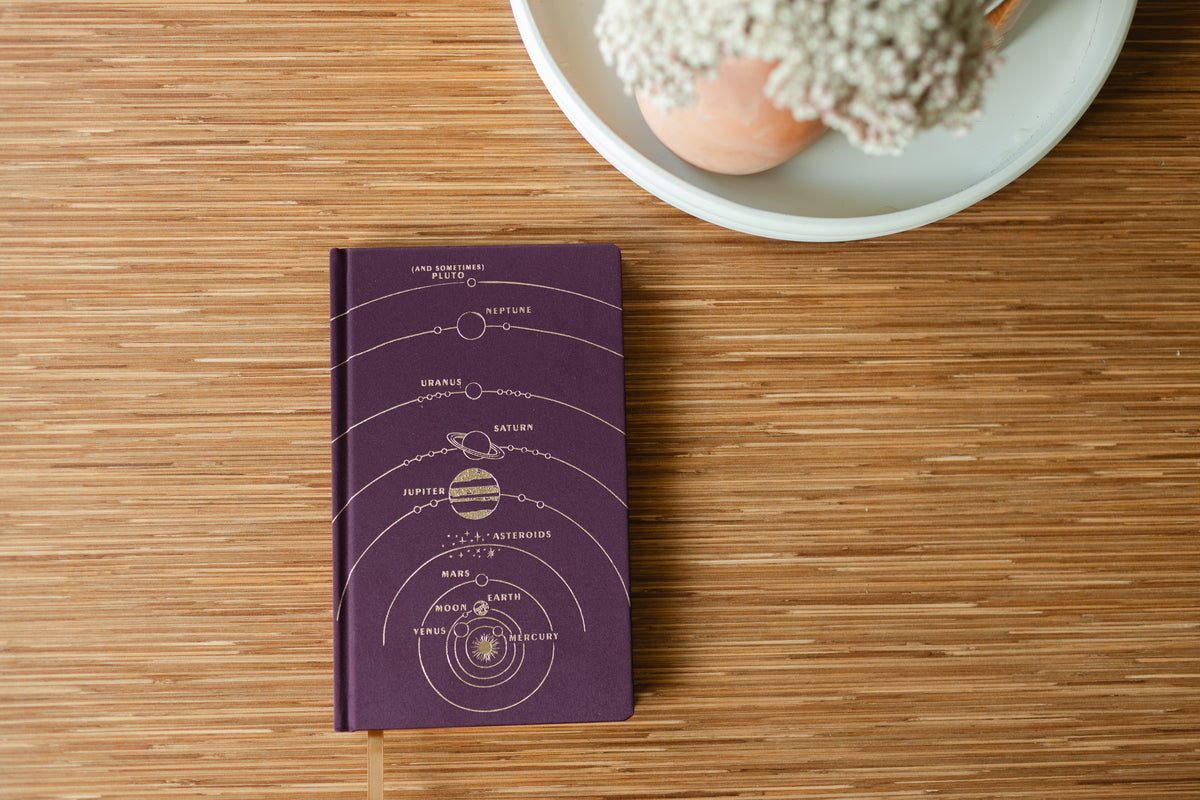 The Cloth Journal - Solar System, featuring a purple cover with lay-flat binding and a gold solar system illustration, rests on a wooden surface beside a white plate with a vase of small white flowers.