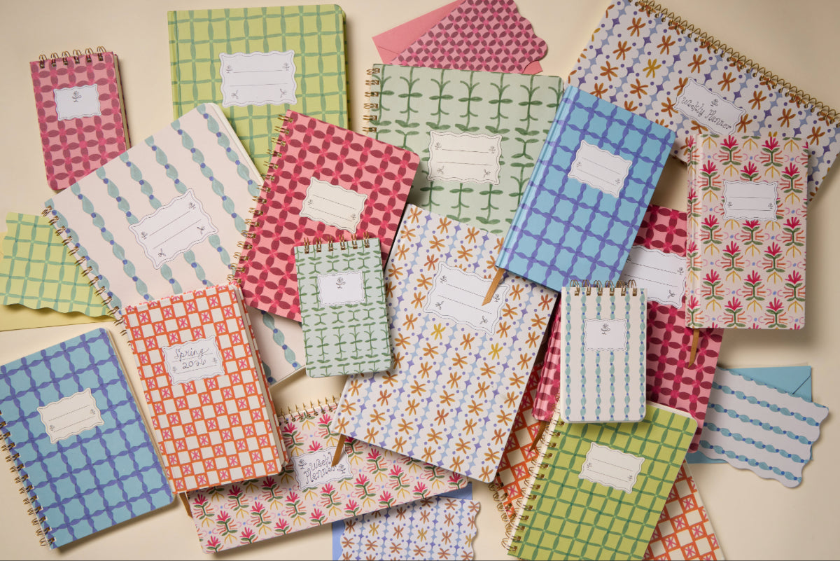 A selection of colorful notebooks and notepads, such as the Tulip Twin Wire Notepad - Green Vine with floral and geometric designs, displayed on a light surface. Some covers offer labeled spaces for names or subjects.