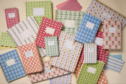 A selection of colorful notebooks and notepads, such as the Tulip Twin Wire Notepad - Green Vine with floral and geometric designs, displayed on a light surface. Some covers offer labeled spaces for names or subjects.