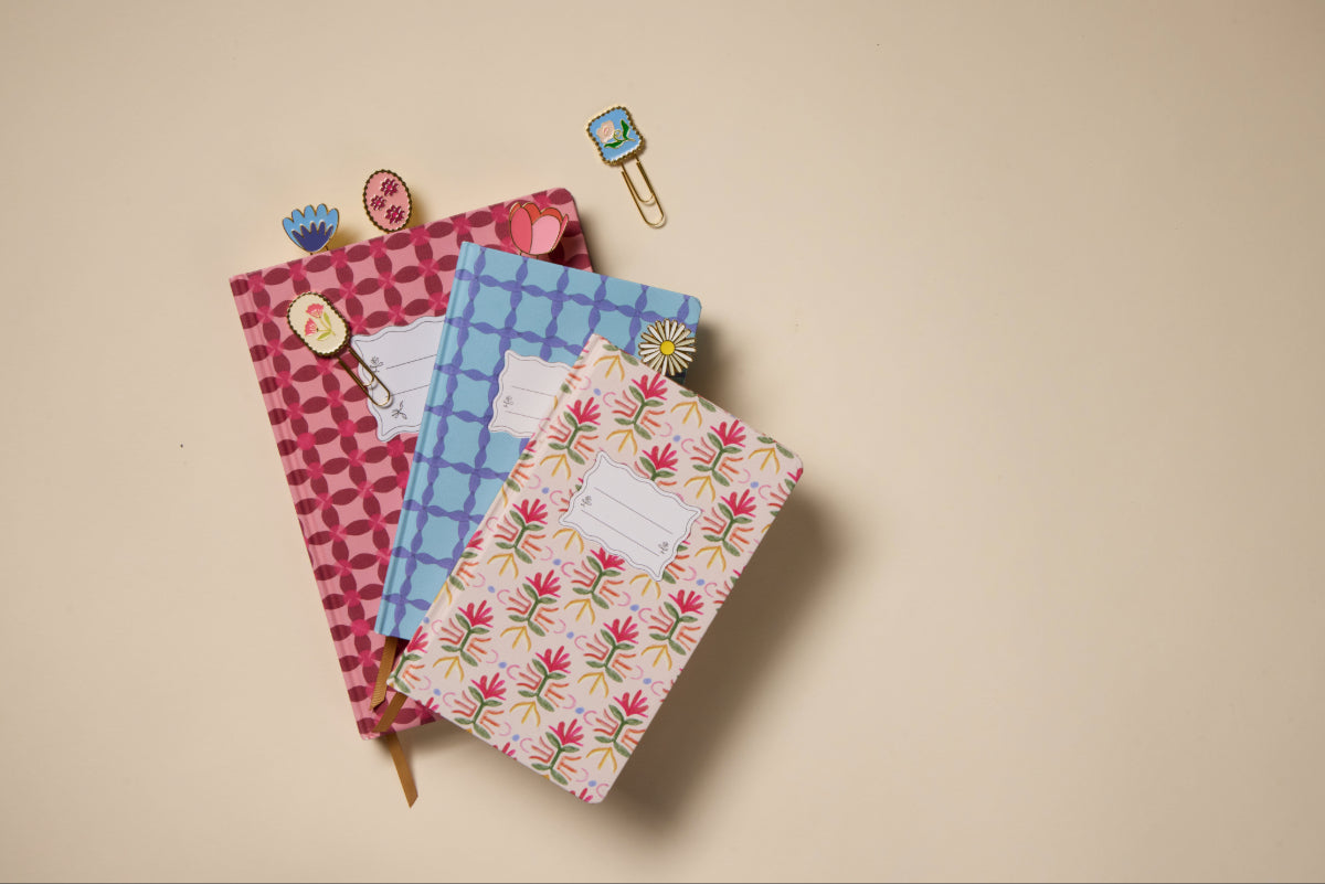 The Tulip Journal - Tropical featuring a vibrant floral cover is stacked on a beige surface, accompanied by decorative paper clips with floral and animal motifs clipped to the journal and scattered nearby.