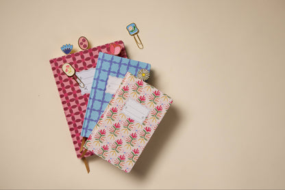 The Tulip Journal - Tropical featuring a vibrant floral cover is stacked on a beige surface, accompanied by decorative paper clips with floral and animal motifs clipped to the journal and scattered nearby.